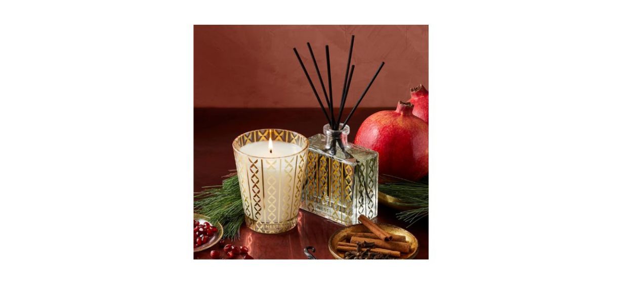 NEST Fragrances Reed Diffuser sitting on a table with a candle, spices, a pomegranate and a pine branch